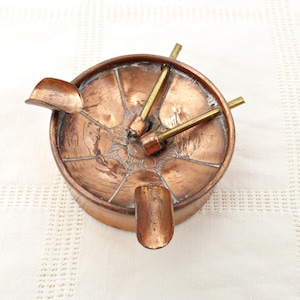 May include: A copper ashtray with four arms that extend out from the center. The arms have a decorative design and are used to hold cigarettes. The ashtray is in good condition with some signs of wear.
