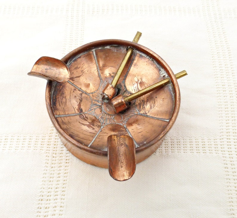 May include: A copper ashtray with four arms that extend out from the center. The arms have a decorative design and are used to hold cigarettes. The ashtray is in good condition with some signs of wear.