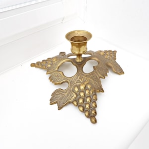 May include: A brass candlestick holder with a grape leaf and cluster design. The candle holder is made of brass and has a round cup to hold a single candle. The base is shaped like a grape leaf with a cluster of grapes.