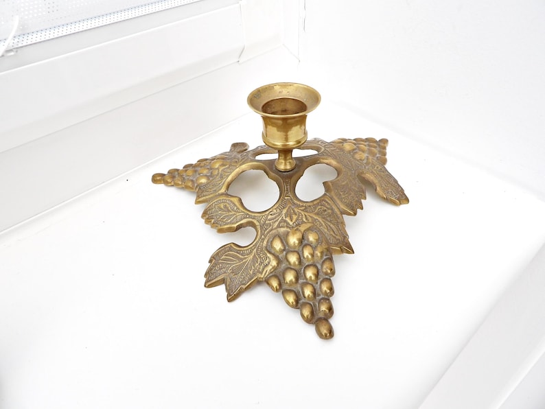 May include: A brass candlestick holder with a grape leaf and cluster design. The candle holder is made of brass and has a round cup to hold a single candle. The base is shaped like a grape leaf with a cluster of grapes.