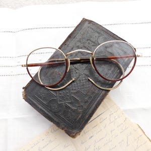 Puede incluir: Gafas redondas vintage con montura dorada y detalles burdeos sobre un libro antiguo de cuero repujado oscuro. Las gafas tienen un dise&ntilde;o cl&aacute;sico, atemporal, que sugiere una est&eacute;tica retro o antigua. La escena evoca nostalgia e intelectualismo.