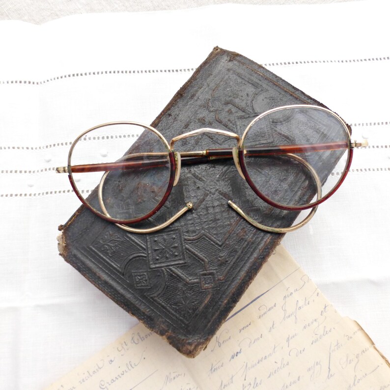 Puede incluir: Gafas redondas vintage con montura dorada y detalles burdeos sobre un libro antiguo de cuero repujado oscuro. Las gafas tienen un dise&ntilde;o cl&aacute;sico, atemporal, que sugiere una est&eacute;tica retro o antigua. La escena evoca nostalgia e intelectualismo.