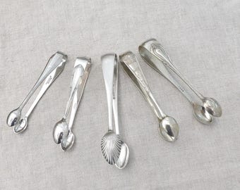 May include: Five silver sugar tongs with various handle designs and spoon-shaped ends are arranged on a neutral-colored surface. The tongs are made of shiny metal, reflecting light, and are likely vintage or antique.