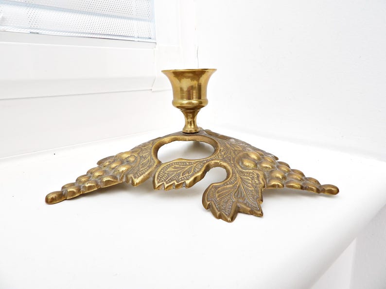 May include: A vintage brass candle holder with a leaf-shaped base. The candle holder has a detailed design with a central cup for the candle and a decorative leaf pattern. The brass has a warm, aged patina.