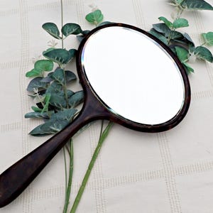 Op de afbeelding: Ovale handspiegel met een donkerbruin, schildpadpatroon frame en handvat. De spiegel heeft een decoratieve rand. De handgreep is lang en slank. De spiegel ligt op een wit oppervlak met groen blad.