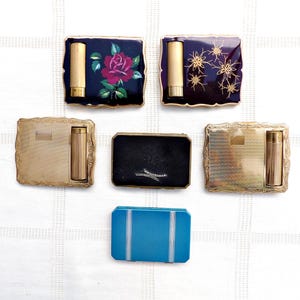 May include: A collection of six rectangular vanity cases and compacts. Two feature dark blue enamel with floral designs, two are gold-toned metal with textured surfaces, one is a black case with a silver accent, and one is a bright blue case.