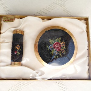 Peut inclure: Un ensemble composé d'un étui à rouge à lèvres et d'un poudrier rond doré, tous deux recouverts de tissu noir avec une broderie florale multicolore. Les objets sont présentés dans une boîte rectangulaire doublée de tissu crème, marquée madaer.