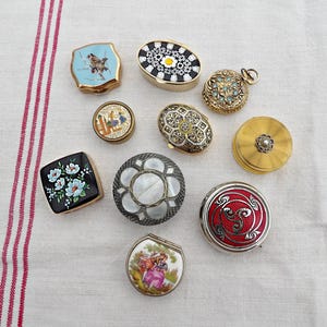 May include: A collection of decorative vintage trinket boxes in various shapes and designs. The boxes feature intricate patterns, including floral, geometric, and mosaic styles. Colors include gold, black, white, blue, and red. Some boxes have hinged lids.