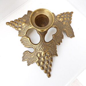 May include: A vintage brass candle holder with a grape and leaf design. The candle holder has a round cup for the candle and is shaped like a grape vine. The brass has a warm, aged patina.