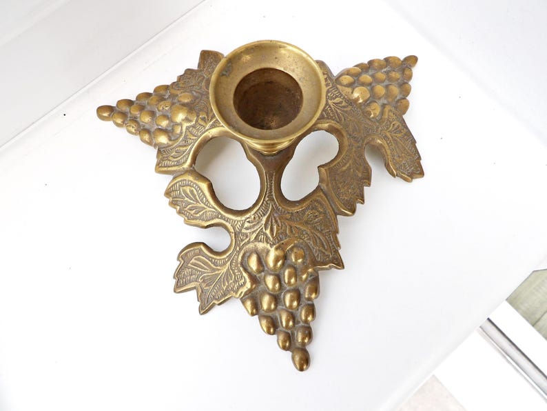 May include: A vintage brass candle holder with a grape and leaf design. The candle holder has a round cup for the candle and is shaped like a grape vine. The brass has a warm, aged patina.