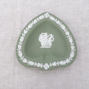 May include: A heart-shaped, sage green ashtray with a white floral and leaf border. The centre features a white relief of classical figures. A small notch is at the bottom. Decorative home decor.