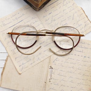 Puede incluir: Gafas redondas vintage con montura de metal dorado y detalles en marr&oacute;n y burdeos. Las gafas descansan sobre cartas manuscritas antiguas, evocando una sensaci&oacute;n de historia y nostalgia. Las lentes son claras.