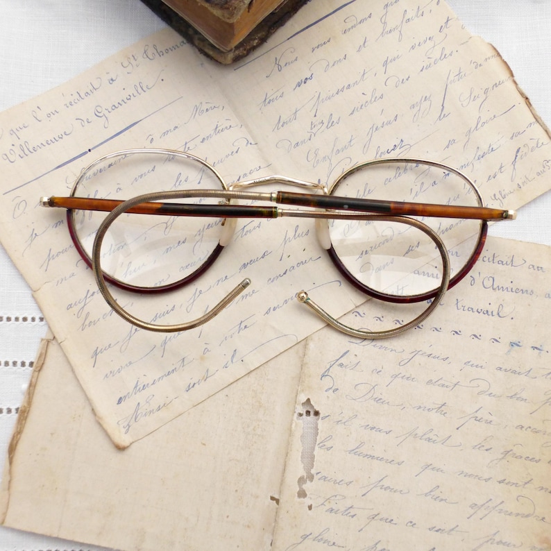 Puede incluir: Gafas redondas vintage con montura de metal dorado y detalles en marr&oacute;n y burdeos. Las gafas descansan sobre cartas manuscritas antiguas, evocando una sensaci&oacute;n de historia y nostalgia. Las lentes son claras.