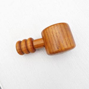 Könnte beinhalten: Ein Holzhammer mit zylindrischem Kopf und einem Griff mit drei abgerundeten Ringen. Der Hammer ist aus hellbraunem Holz gefertigt und vor einem weißen Hintergrund platziert. Die Holzmaserung ist sichtbar.