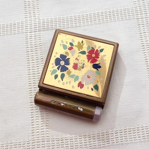 May include: A vintage compact mirror with a gold-toned metal frame. The top features a floral design in red, blue, and white. The compact has a small, cylindrical handle with decorative accents. The background is a white, textured fabric.