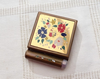 May include: A vintage compact mirror with a gold-toned metal frame. The top features a floral design in red, blue, and white. The compact has a small, cylindrical handle with decorative accents. The background is a white, textured fabric.