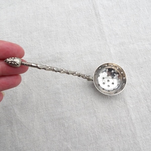 Peut inclure: Une passoire à thé en métal argenté dotée d'un long manche fin orné d'un motif texturé en forme de feuilles et d'une extrémité arrondie. Le bol circulaire de la passoire est perforé d'un motif répétitif de petites étoiles à six branches.