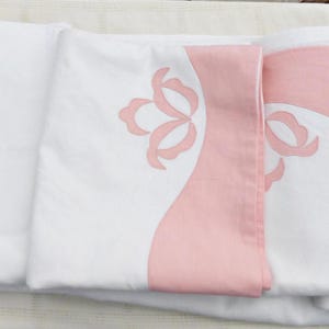 Op de afbeelding: Een set witte linnen servetten met roze geborduurde bloemenmotieven. De servetten zijn gevouwen en gestapeld op een wit oppervlak.