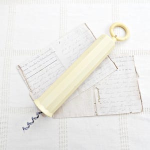 May include: Ivory-colored corkscrew with a ring handle and a spiral metal worm. The corkscrew is lying on top of several antique handwritten letters. The letters are faded and aged, adding to the vintage aesthetic.