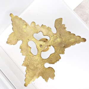 May include: A brass-colored decorative furniture pull with a leaf design. The pull features a detailed, openwork leaf shape with a central knob and screw. The finish has a slightly aged appearance.