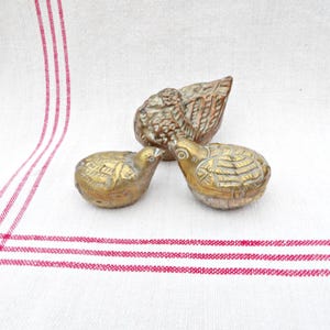 May include: Three antique brass bird figurines. Two small birds are side-by-side, and a larger bird is behind them. The birds have detailed feather and wing designs. The figurines are on a white cloth with red stripes.