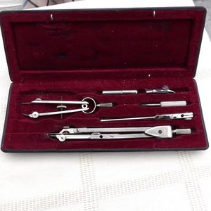 May include: A vintage drafting set in a black case with a burgundy velvet lining. The set includes several silver-colored compasses and dividers, ideal for technical drawing and geometry. The case is open, displaying the tools neatly arranged.