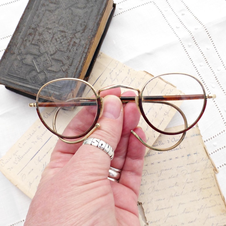 Puede incluir: Gafas redondas vintage con montura dorada y patillas marrones. Las gafas tienen un dise&ntilde;o &uacute;nico con varillas curvas. Tambi&eacute;n se muestra un libro antiguo y cartas manuscritas.