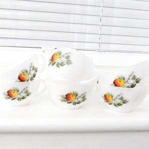 May include: Set of six white ceramic teacups with a floral design featuring apples and leaves. The cups have a handle on the side and are arranged in a row on a white surface.