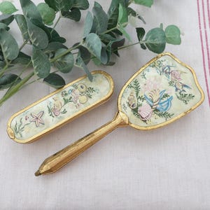 May include: A vintage vanity set featuring a hand mirror and a matching compact. Both pieces have a gold-tone frame and are decorated with a seashell and floral design in pastel colors. The set is displayed on a white surface with a sprig of greenery.