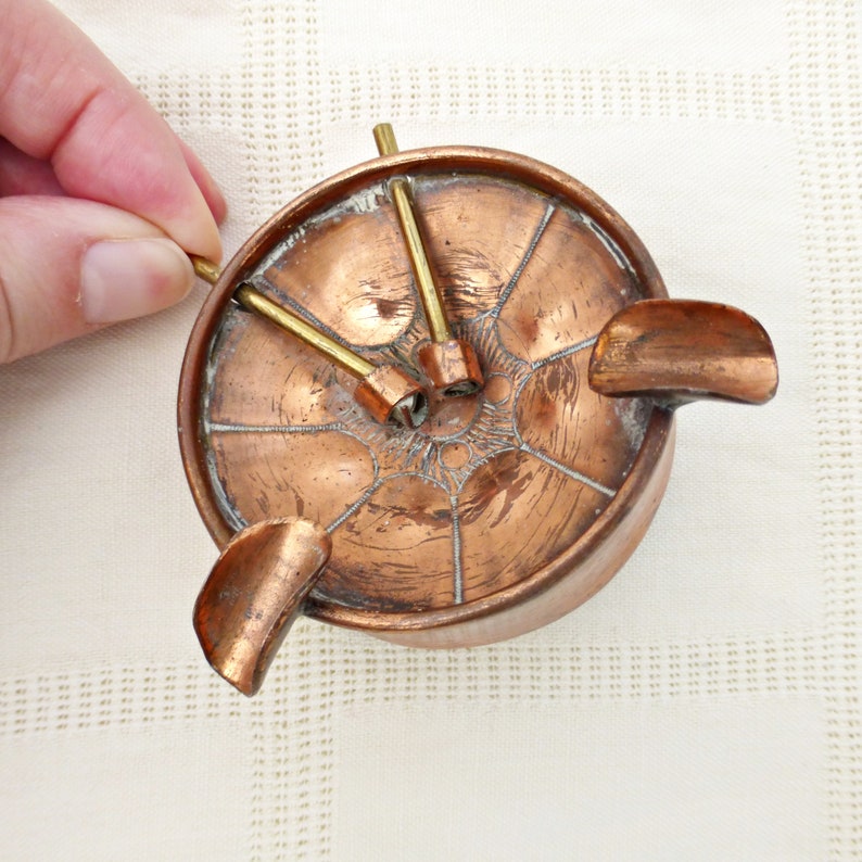 May include: A copper ashtray with three brass rods extending from the center. The ashtray has a decorative design and a raised lip around the edge.