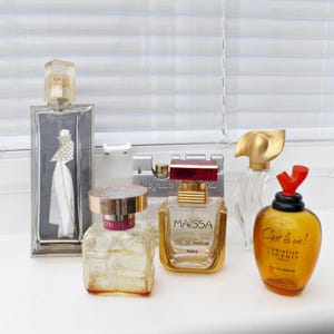 May include: Five different perfume bottles, including a clear bottle with a gold cap and a white label that says "C'est la vie! Christian Lacroix Paris Eau de Parfum", a clear bottle with a gold cap and a pink label that says "Maissa Eau de Parfum Paris", a clear bottle with a gold cap and a white label that says "Reyne", a clear bottle with a gold cap and a white label that says "C'est la vie! Christian Lacroix Paris Eau de Parfum", and a clear bottle with a gold cap and a white label that says "C'est la vie! Christian Lacroix Paris Eau de Parfum".