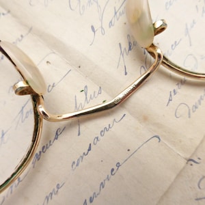 Puede incluir: Primer plano de gafas doradas vintage con lentes redondas. Las monturas tienen un dise&ntilde;o delicado, con puente y varillas. Las lentes se sujetan con peque&ntilde;os soportes curvos. Las gafas descansan sobre un fondo de escritura manuscrita.
