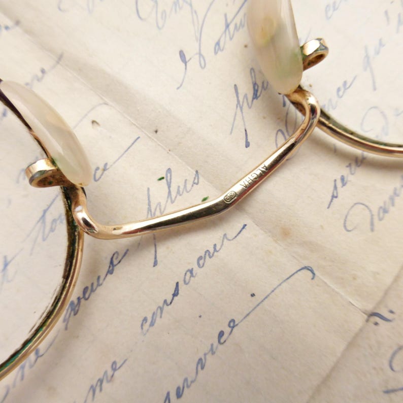 Puede incluir: Primer plano de gafas doradas vintage con lentes redondas. Las monturas tienen un dise&ntilde;o delicado, con puente y varillas. Las lentes se sujetan con peque&ntilde;os soportes curvos. Las gafas descansan sobre un fondo de escritura manuscrita.