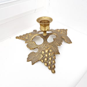 May include: A brass candle holder with a grape leaf design. The candle holder has a round cup for the candle and is attached to a detailed leaf shape with a cluster of grapes. The brass has a warm, antique finish.