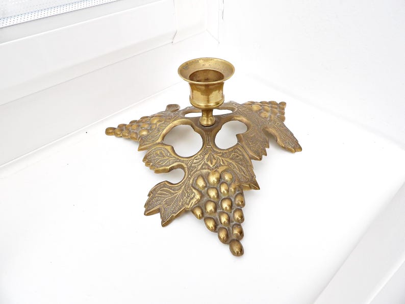 May include: A brass candle holder with a grape leaf design. The candle holder has a round cup for the candle and is attached to a detailed leaf shape with a cluster of grapes. The brass has a warm, antique finish.