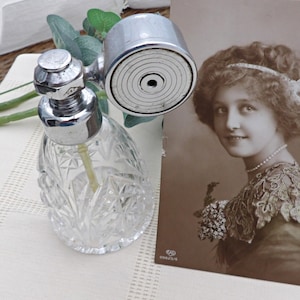 May include: A vintage perfume bottle with a clear, cut-glass body and a silver-toned atomizer. The bottle is next to a sepia-toned portrait of a woman. The atomizer has a circular spray head.