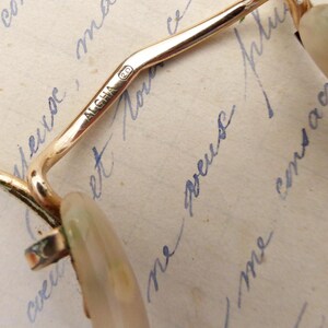 Puede incluir: Primer plano de gafas vintage doradas con lentes ovaladas. La montura presenta la inscripci&oacute;n "ALGHA 20". Las lentes tienen un tono verde claro y beige. Las gafas est&aacute;n sobre un fondo de escritura manuscrita.