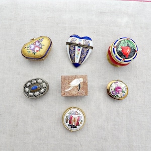 May include: A collection of seven small decorative trinket boxes featuring various shapes and designs, including a gold heart with floral patterns, a blue and white heart, a round red box with a flower, a silver oval box, a rectangular stone box with a bird, a round floral box, and a round butterfly box.