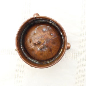 May include: A copper-colored metal container with a round top and three legs. The container has a slightly dented surface and a small opening in the top.