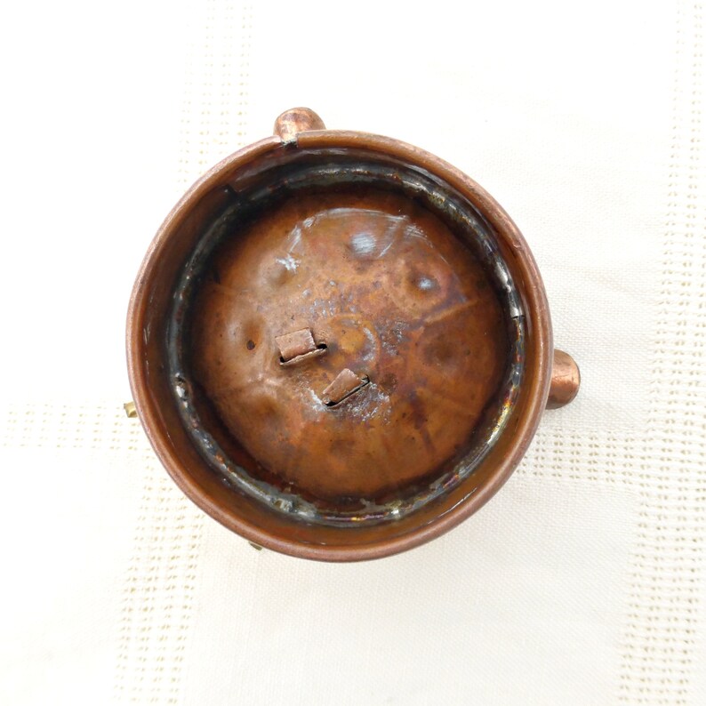 May include: A copper-colored metal container with a round top and three legs. The container has a slightly dented surface and a small opening in the top.