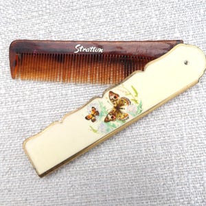 May include: A folding pocket comb with a brown tortoiseshell-patterned plastic comb section featuring the white text Stratton. The handle is an off-white rectangular case with a scalloped edge, decorated with a colorful butterfly illustration.