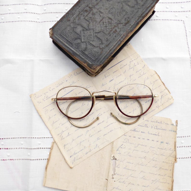 Puede incluir: Gafas redondas vintage con montura dorada y detalles marrones sobre cartas manuscritas antiguas. Un libro antiguo con una cubierta oscura y texturizada tambi&eacute;n est&aacute; presente. Los objetos est&aacute;n sobre una superficie blanca texturizada.