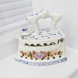 May include: A white ceramic iron with blue and floral designs. The iron has a handle and a hinged lid. The iron is a vintage decorative piece.