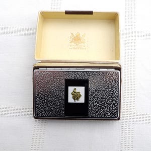 Op de afbeelding: Een open, vintage sigarettenetui met een getextureerde, zilverkleurige buitenkant en een zwart middenpaneel. De etui zit in een crèmekleurige doos met een bruine rand. Een gouden embleem is zichtbaar aan de binnenkant van de doos.