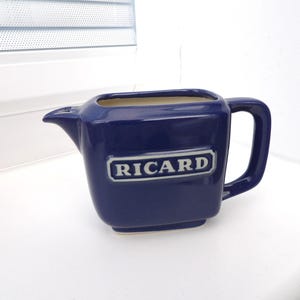 Puede incluir: Una jarra de cerámica azul marino con cuerpo rectangular y asa. La palabra "RICARD" se muestra en letras blancas en la parte delantera. La jarra tiene un pico.