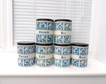 May include: A set of seven ceramic canisters with black lids. The canisters are white with blue decorative panels and text that reads "Chicoree", "Riz", "Farine", "Café", "Sel", and "Sucre".