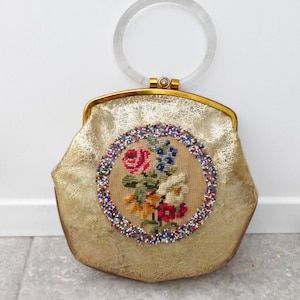 Puede incluir: Un bolso vintage dorado con un asa circular transparente. El bolso presenta un diseño floral bordado con flores rojas, azules y blancas, rodeado por un borde de cuentas. El marco del bolso es dorado.