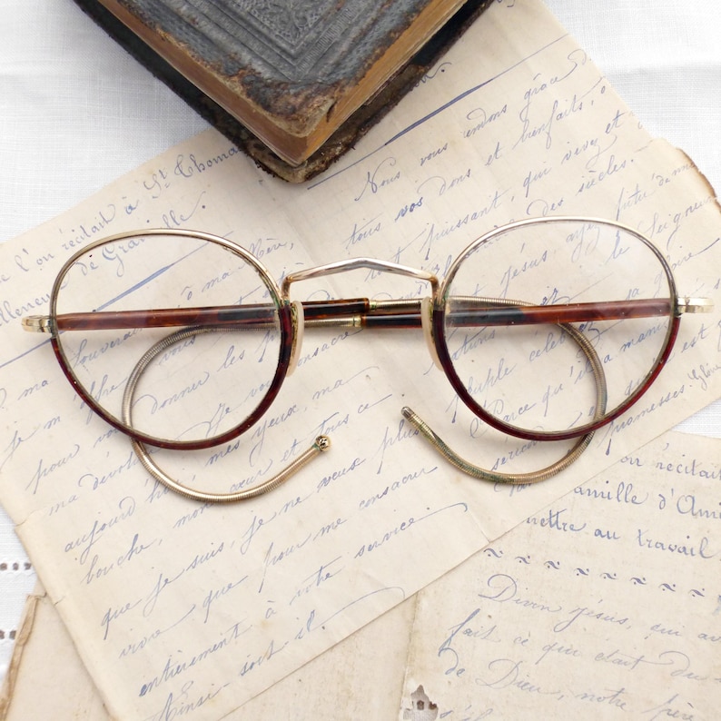 Puede incluir: Gafas redondas vintage con montura dorada y patillas marrones, sobre papel envejecido con escritura manuscrita. Las gafas tienen un dise&ntilde;o cl&aacute;sico y retro, evocando una est&eacute;tica hist&oacute;rica.