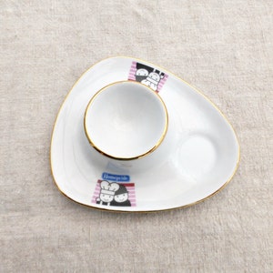 Peut inclure: Un œuf en céramique blanc avec un bord doré et une petite soucoupe. La soucoupe présente une illustration de deux chefs en dessin animé avec le texte "Homepride" dessus.