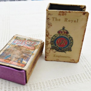Könnte beinhalten: Zwei antike Streichholzschachteln. Eine ist eine rot-weiß-blaue Streichholzschachtel mit dem Text "Pilot Match" und "British Made". Die andere ist eine weiße Streichholzschachtel mit dem Text "The Royal Engineers" und einem rot-weiß-blauen Wappen.